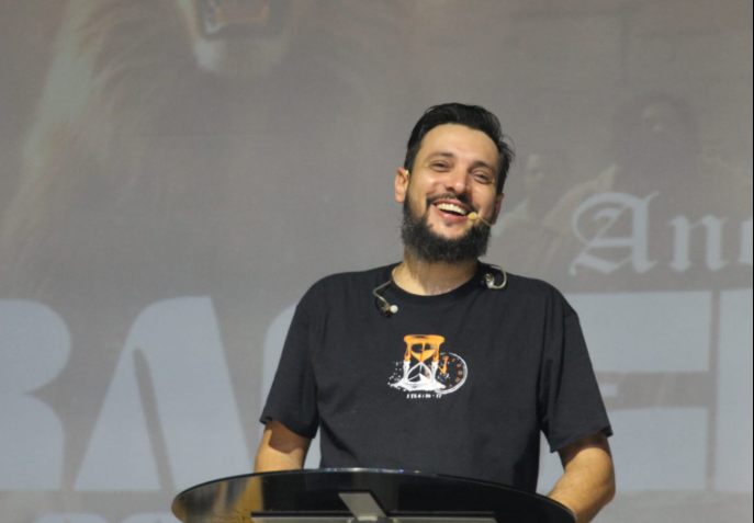 Pastor Felipe Golim em oração, com mãos postas e expressão séria, em palco com iluminação suave, representando sua nova fase