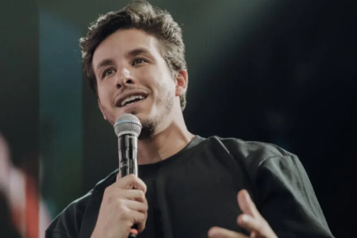 Ricky Tavares em palco de igreja com mãos levantadas em oração, segurando Bíblia, jovens ao fundo.