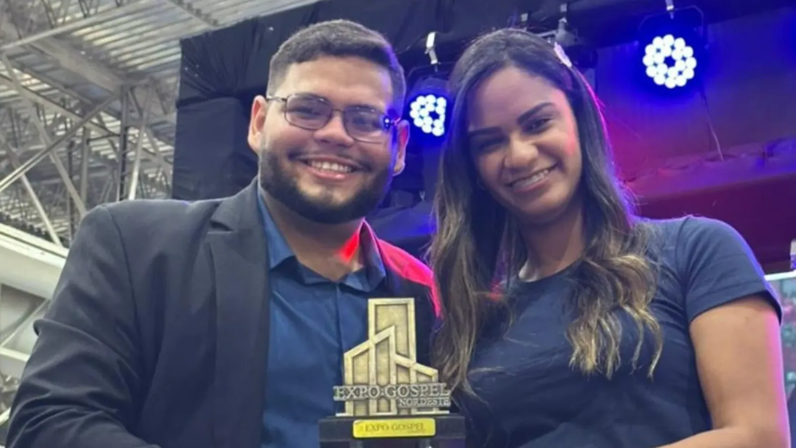 Cantor Davi Sacer se apresenta em palco de evento gospel com microfone e banda, sob luzes coloridas, em ginásio lotado.