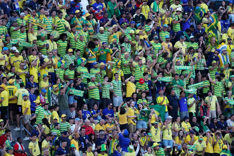 Torcedores brasileiros no estádio da Copa do Mundo, alguns com camisas da seleção e outros com faixas religiosas, em clima de